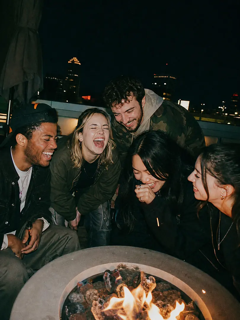 Friends laughing around a fire pit on a city rooftop at night
